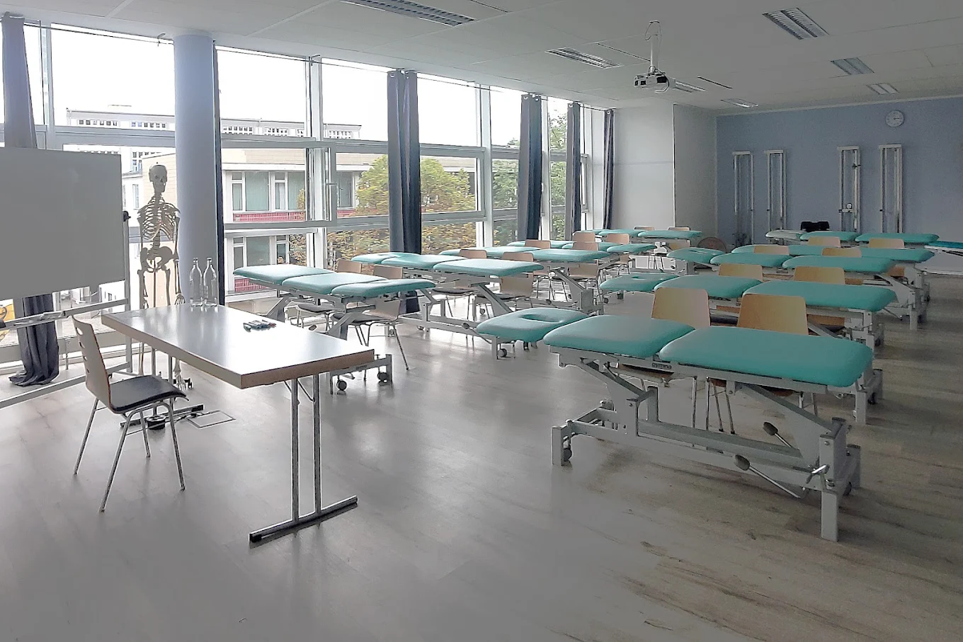 TOP-PHYSIO Seminarraum mit White Board, Dozententisch mit Stuh, Behandlungsliegen und Stühlen und breiter Fensterfront